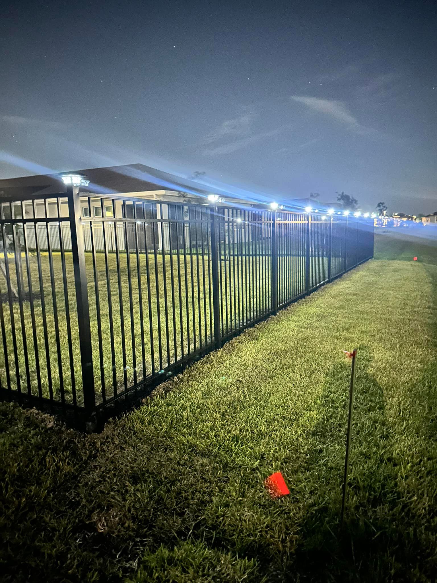 Black metal fence at night, illuminated by lights, in a grassy area. Sky is dark with faint stars.
