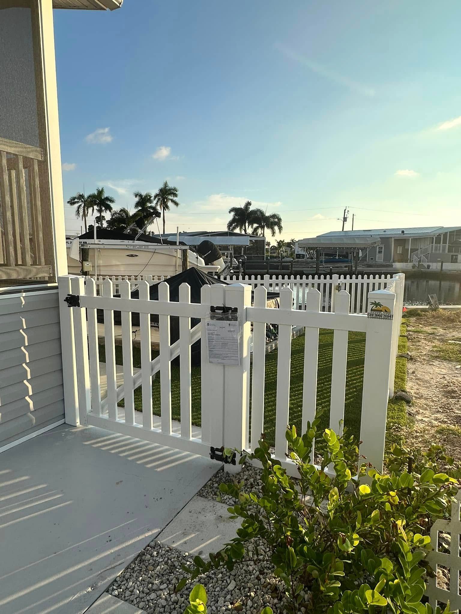 White picket gate on a concrete patio, leading to a yard with a waterfront view; sunny day.