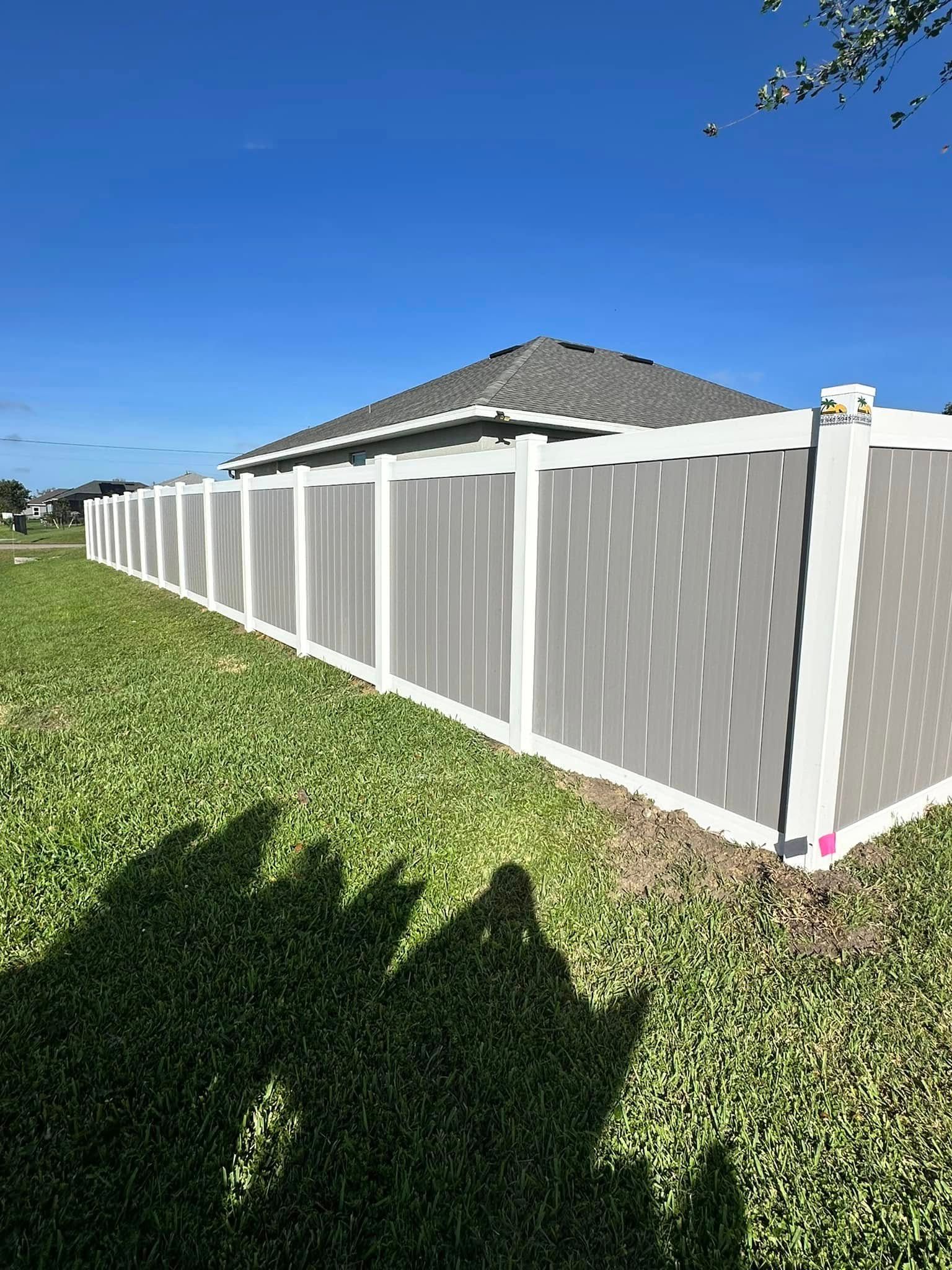 A long white and gray fence surrounds a lawn with a house visible in the background under a blue sky.