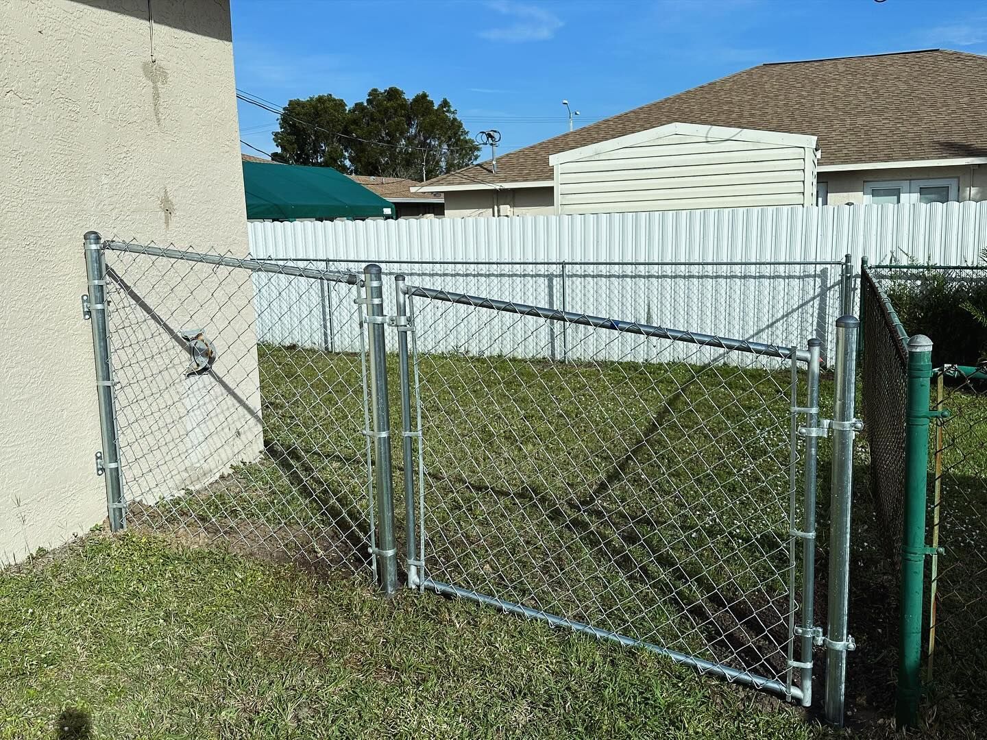 Chain-link fence with two gates in a grassy yard; connecting to a house on one side, and a wooden fence on the other.