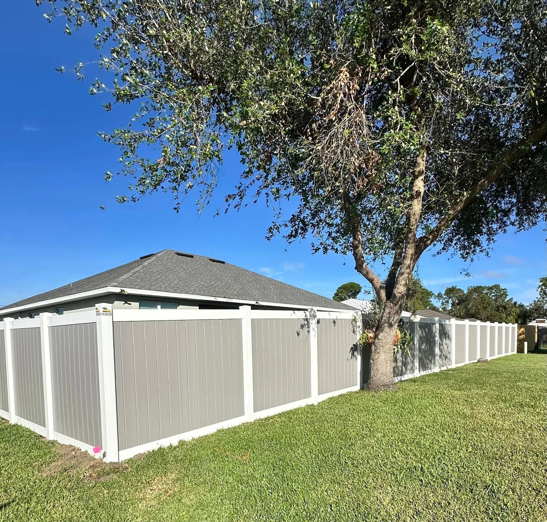 Gray and white fence in front of a house with a tree, on green grass under a blue sky.