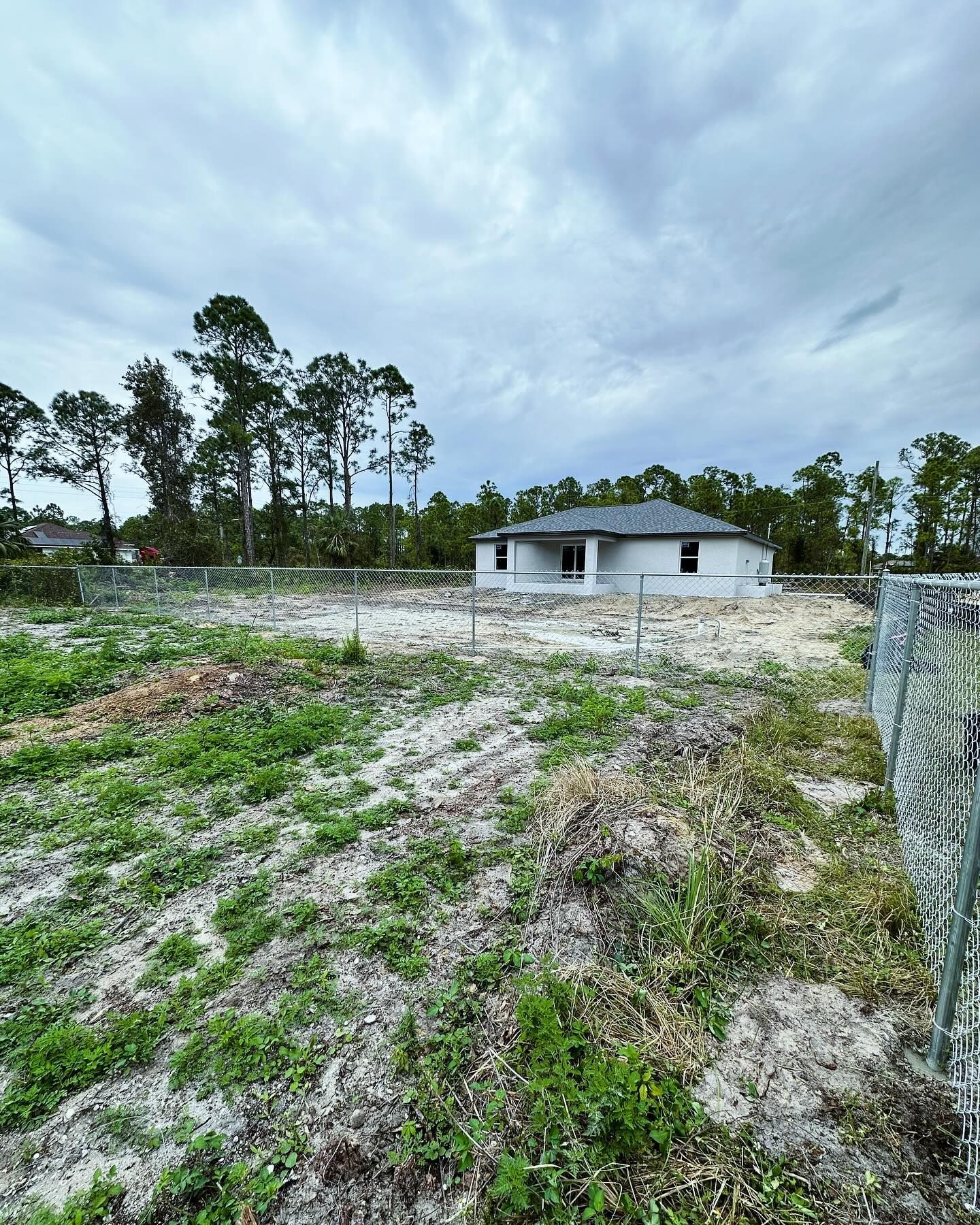 A partially built house on a cleared lot, surrounded by chain-link fence under an overcast sky.
