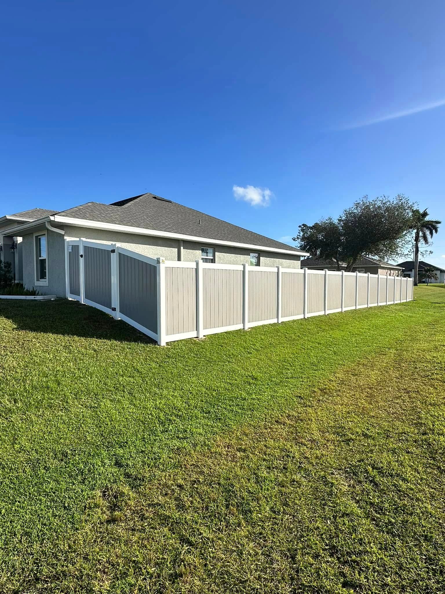 Gray privacy fence surrounds a house with gray roof and green lawn on a sunny day.