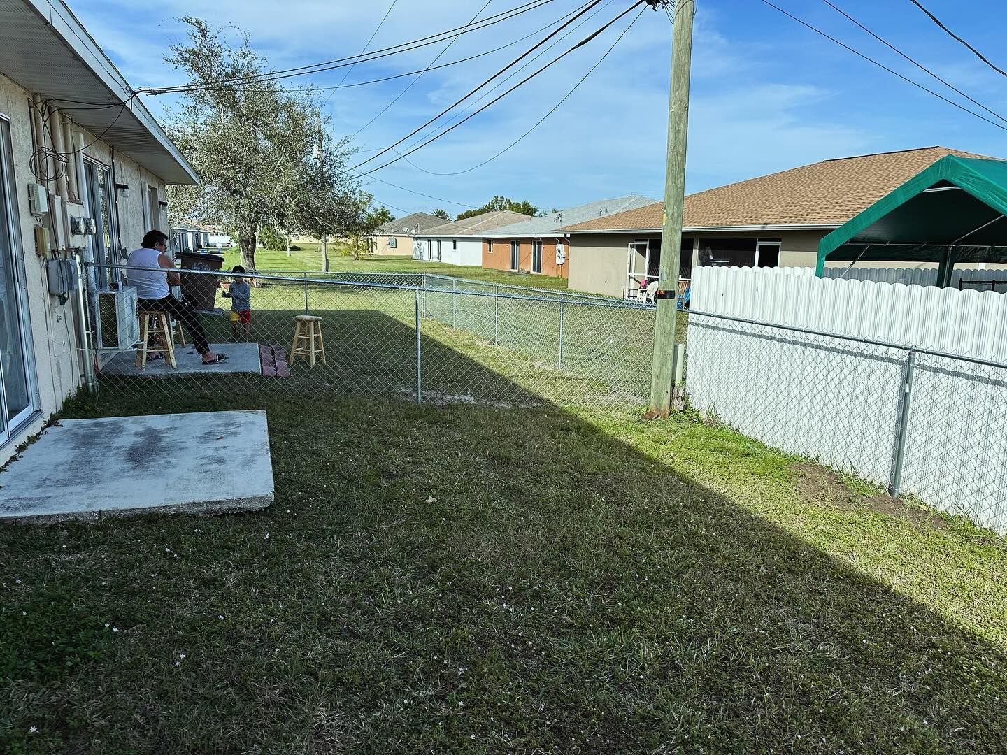 Backyard with a person and child near a house, chain link fence, power lines, sunny day.