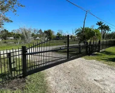 A black fence surrounds a dirt road with palm trees in the background.