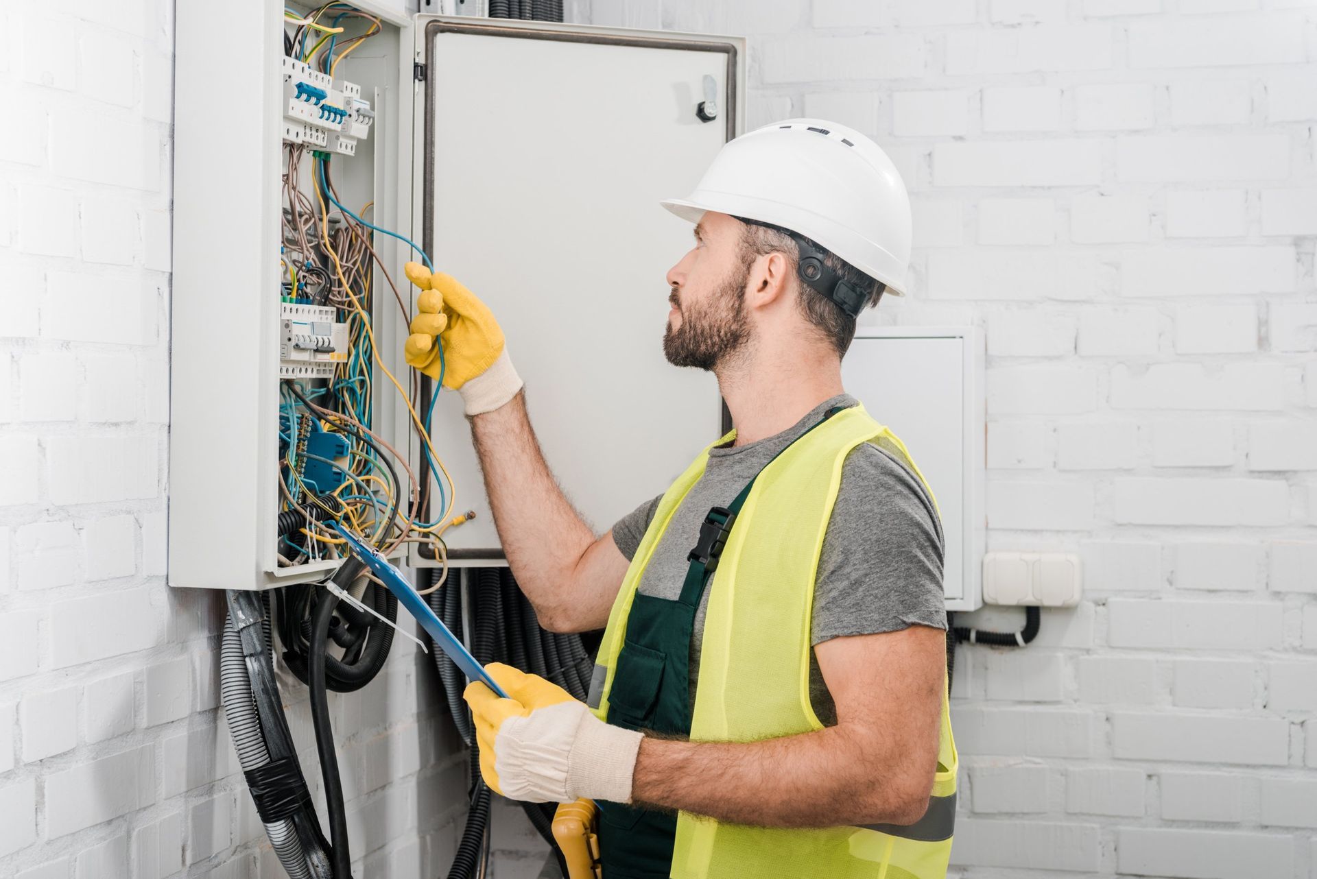 An electrician in a hard hat and high-visibility vest works on an open electrical panel against a white brick wall.