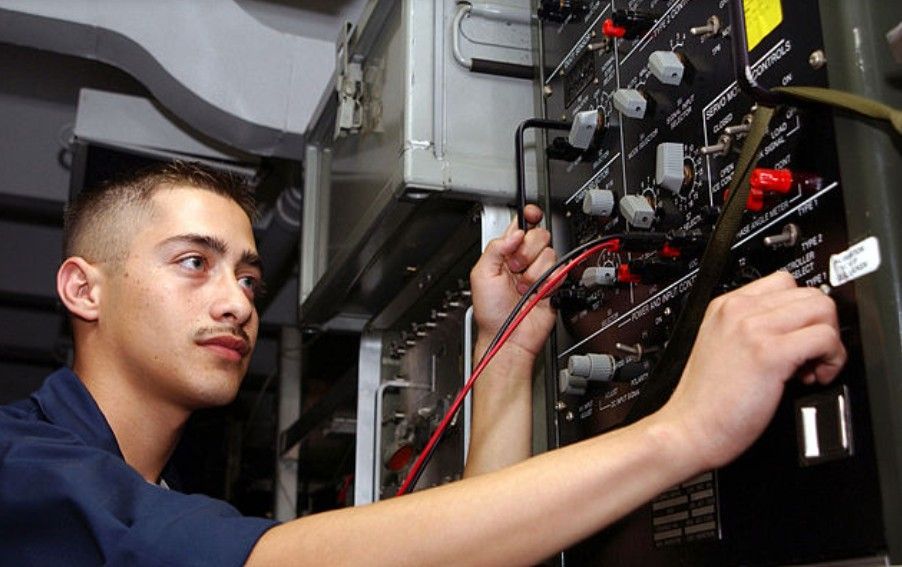 A person wearing work gloves installs a white circular fire alarm sensor into a ceiling grid with exposed wiring.