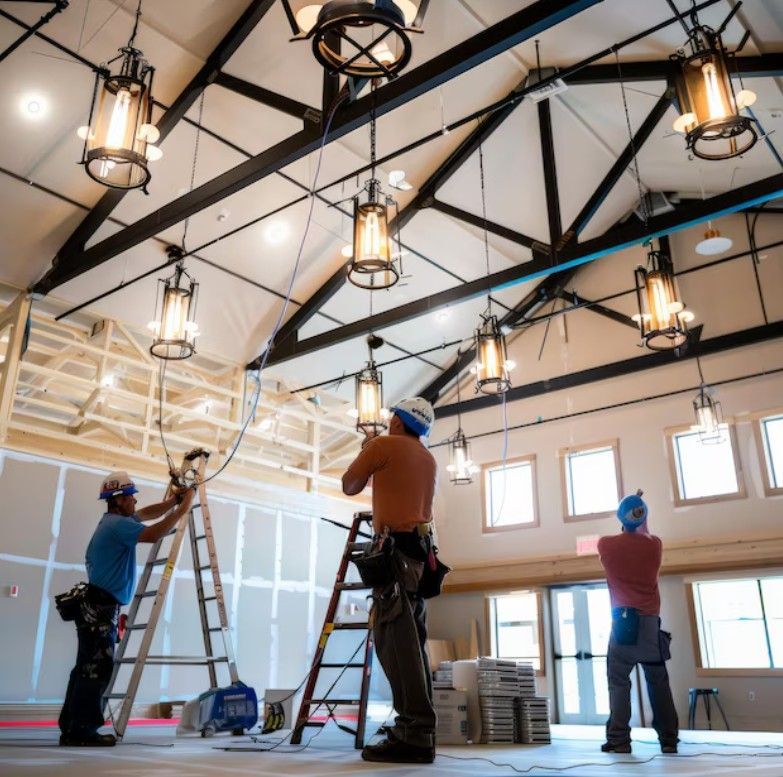 Workers installing hanging lights in a bright industrial space with ladders and exposed beams