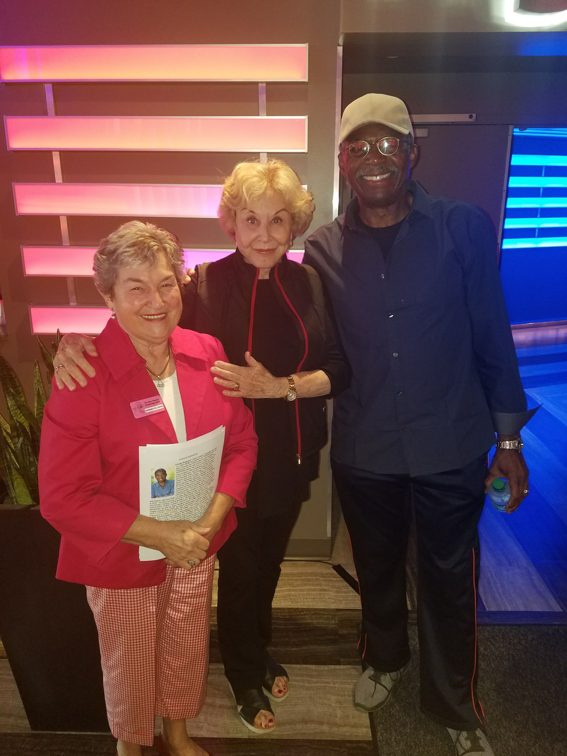 Three people pose for a photo in front of a colorful wall. One person wears red, and another has an arm around one of the other two.