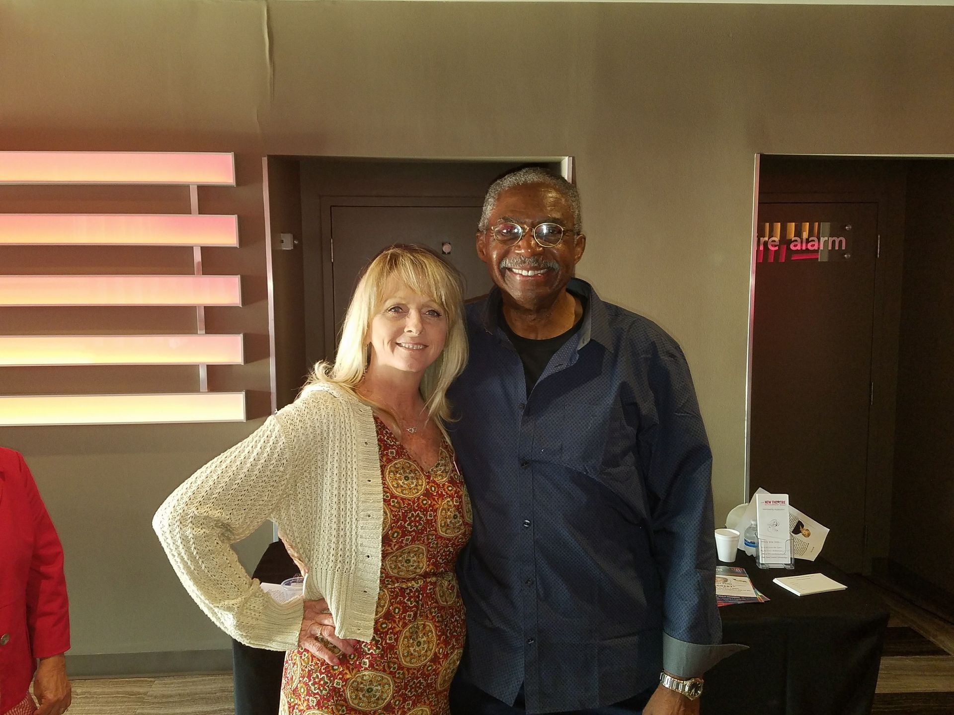 Woman and man smiling, standing near a table and wall with horizontal slats; neutral expressions.
