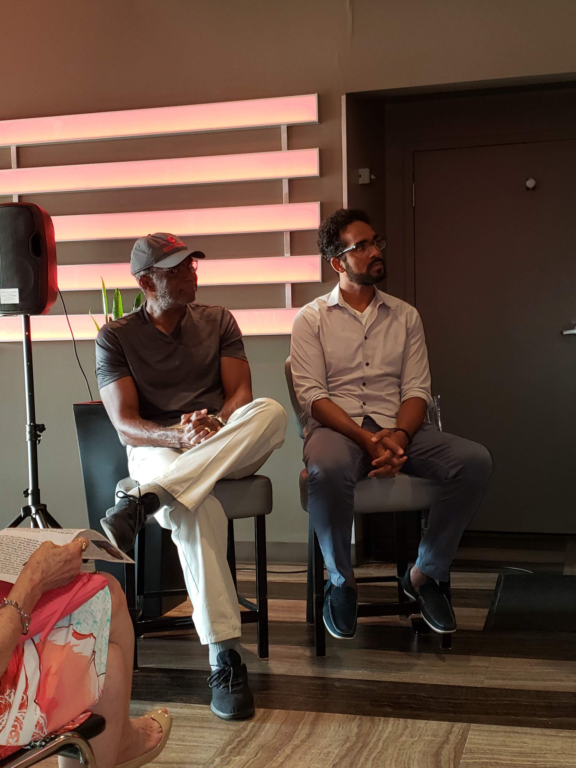 Two men seated on stools; one in cap, grey shirt, cream pants, other in glasses, button-down, jeans, looking forward.