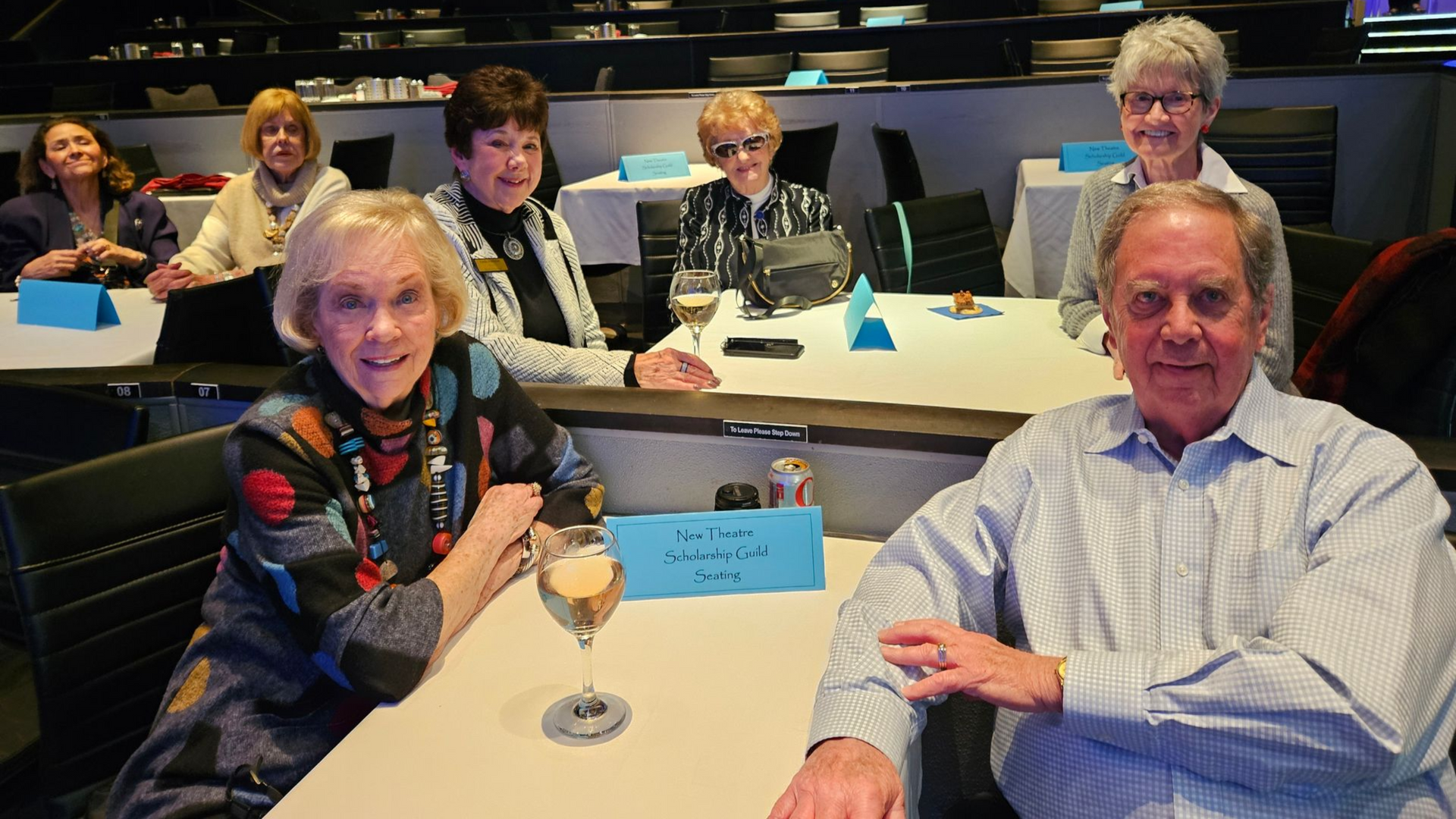 People seated at tables in a venue. Two in the foreground smile; others look towards camera. Tables have drinks and name cards.