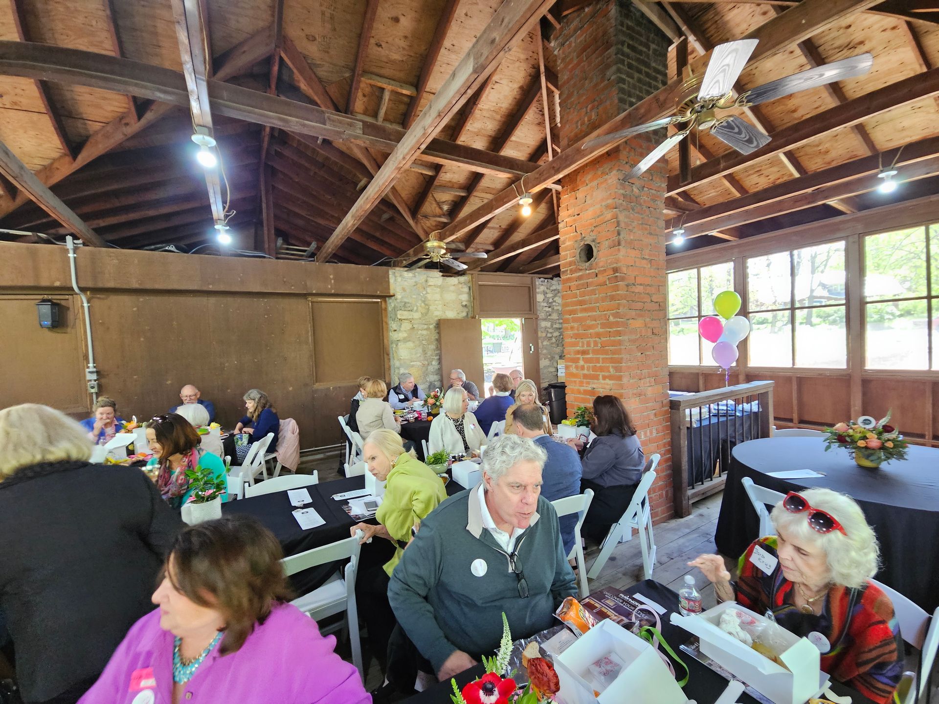 People seated at tables in a pavilion, likely at an event. Floral centerpieces and balloons are visible.