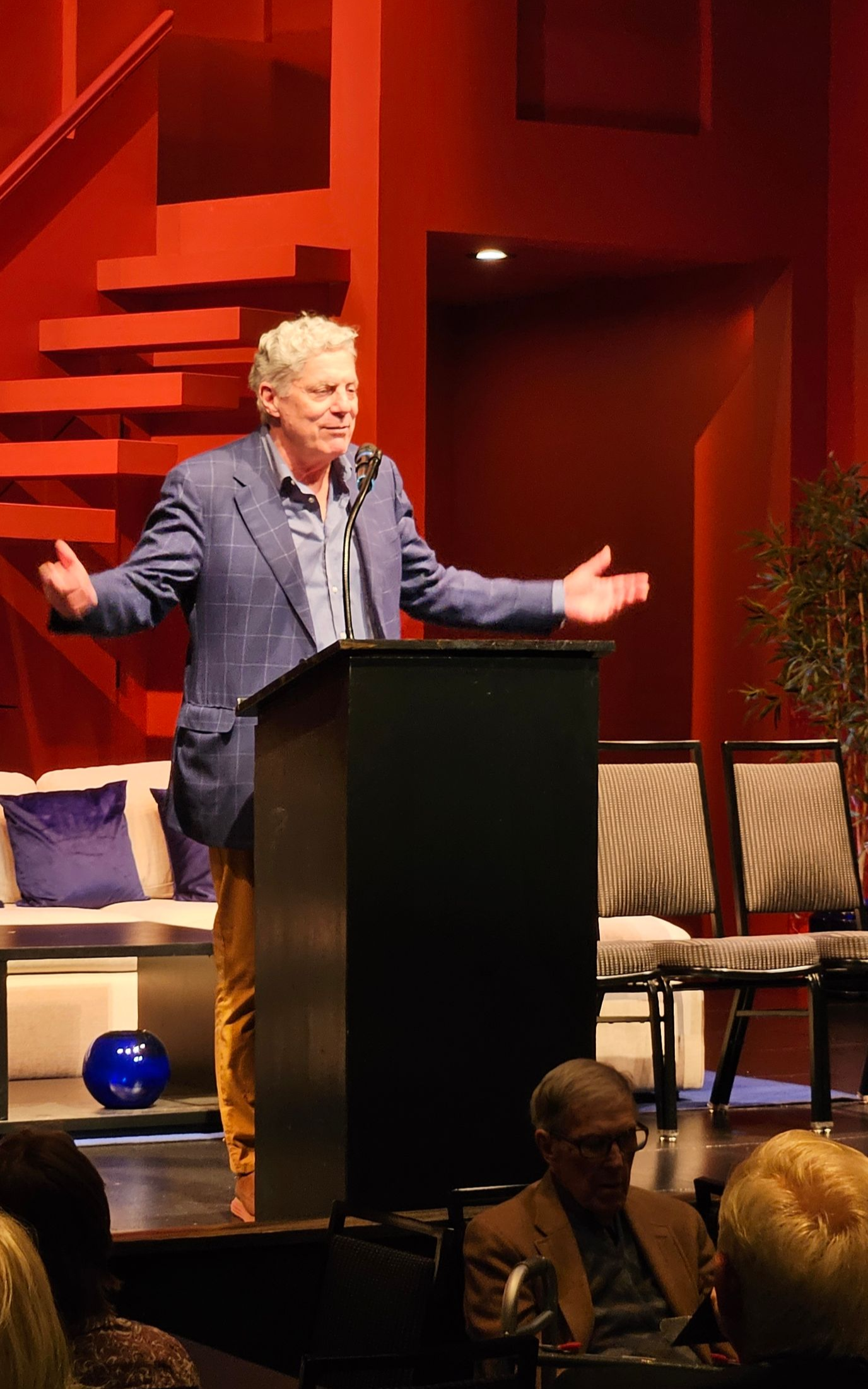 Man speaking at podium with open arms. Red background, chairs, and audience.