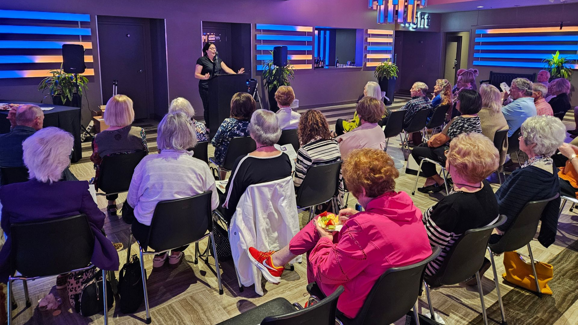 A person speaks to a seated audience in a warmly lit room. Most attendees have white hair.