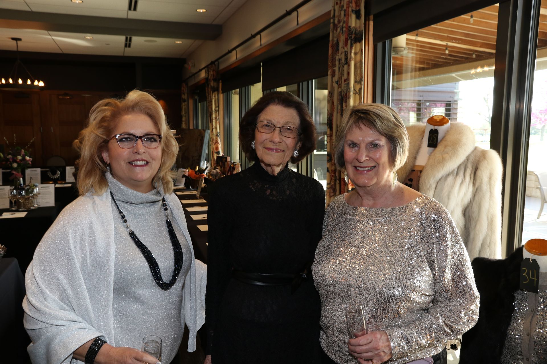 Three women smiling, posing for a photo at an event, near a window and a display with a fur coat.