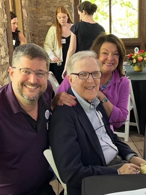 Three people smiling for a photo. A man in glasses is the central figure, flanked by a man on his left and a woman on his right, inside.