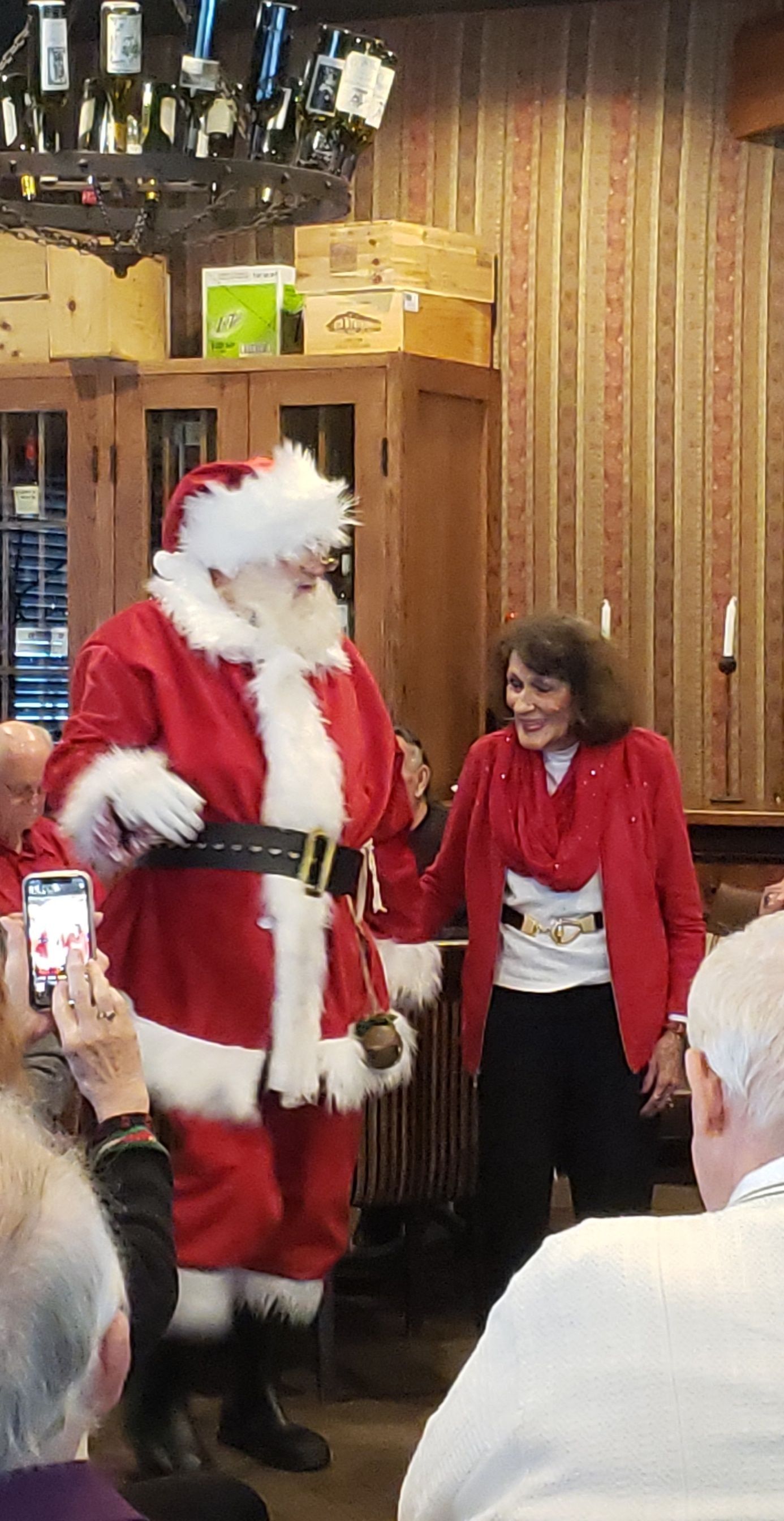 Santa Claus holding hands with a woman in a red jacket, inside a restaurant.