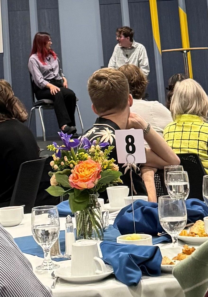 Two people on stage speak at a luncheon. Guests sit at tables with flowers and numbered place settings.