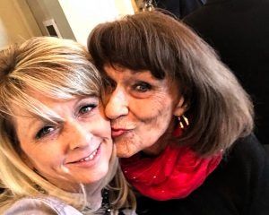 Woman smiles, leans into older woman's cheek, who puckers up wearing a red scarf. Close-up photo.