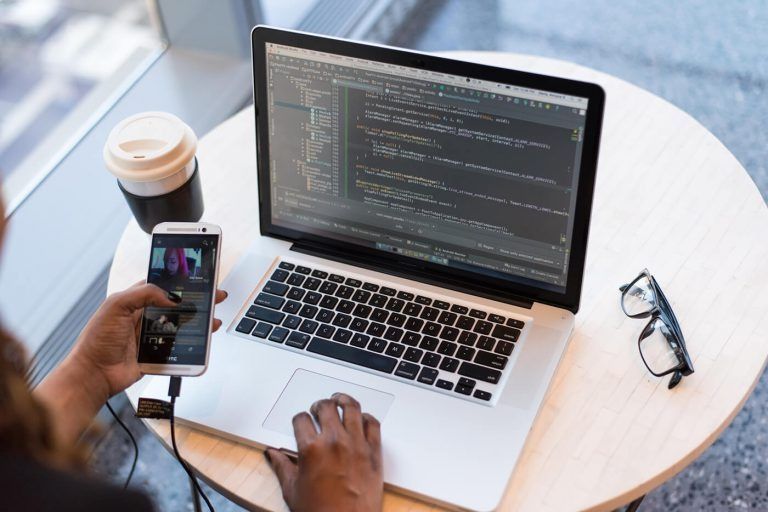 Person using laptop and smartphone with coffee and glasses on table. Coding on the laptop screen.