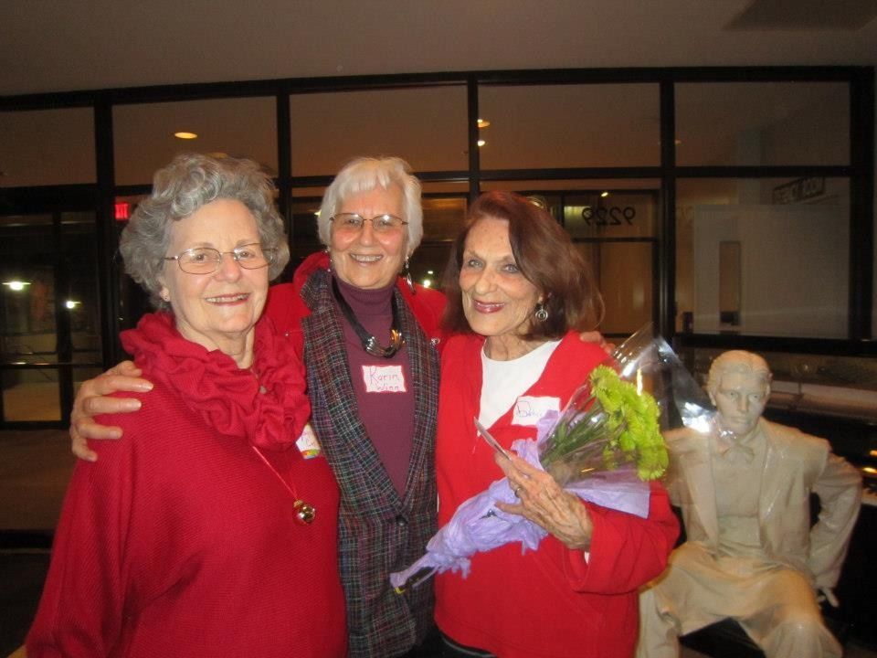 Three women in red coats pose with flowers near a statue outdoors at night.