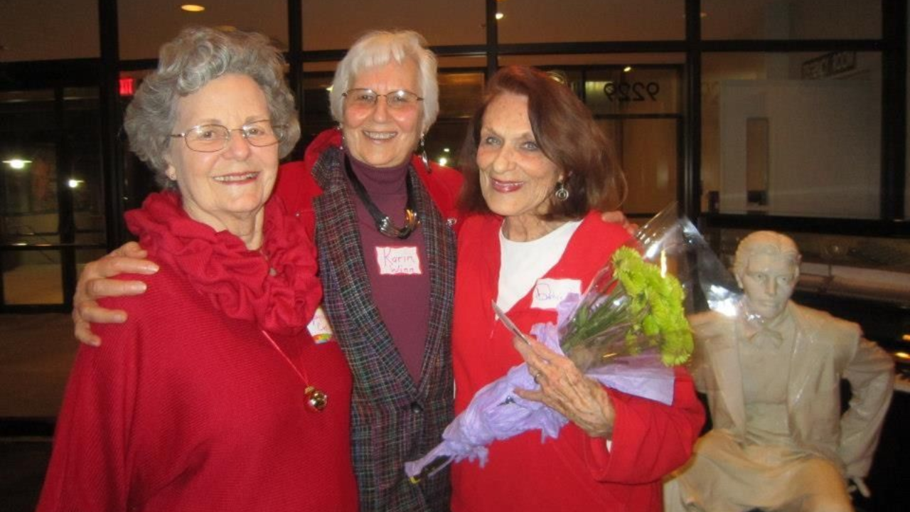 Three people with name tags pose together, one holding flowers, outside a building with statue.