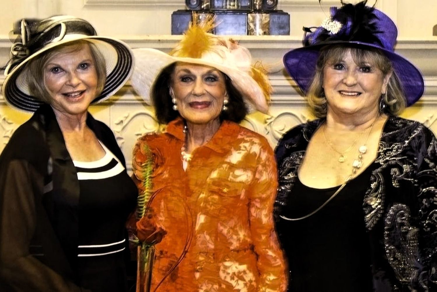 Three women in hats, smiling at a formal event, against a wall with ornate details.