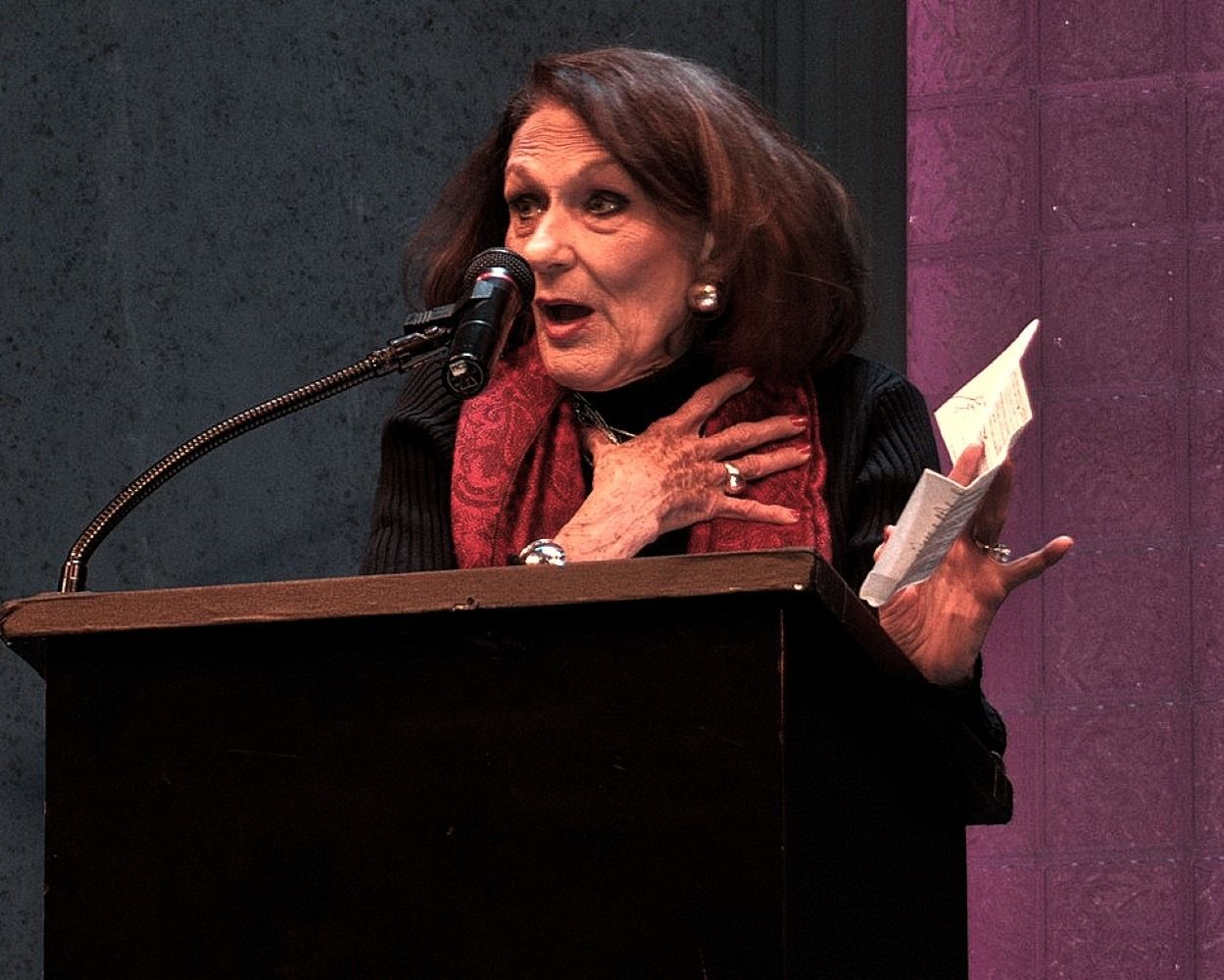 Woman speaking at a podium, holding papers, with an open mouth. She wears a scarf and gestures.
