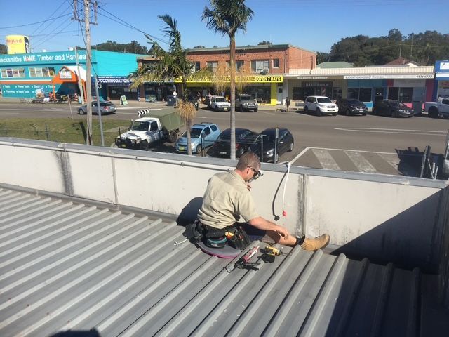 A worker sits on a corrugated metal roof, using a power tool to perform repairs — Channel Electrical In Swansea Heads, NSW