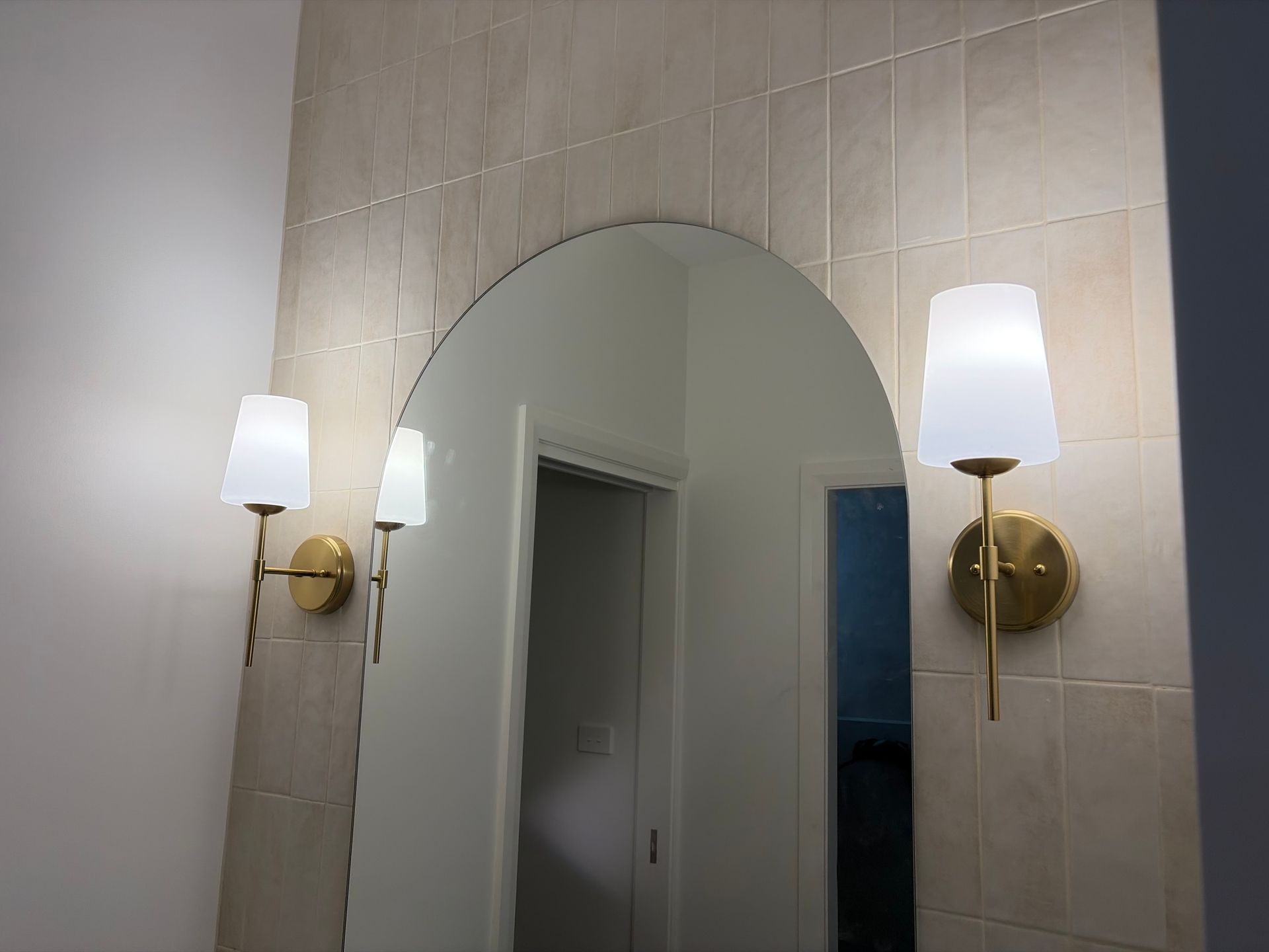 An arched wall mirror centered between two lit brass wall sconces — Channel Electrical In Swansea Heads, NSW