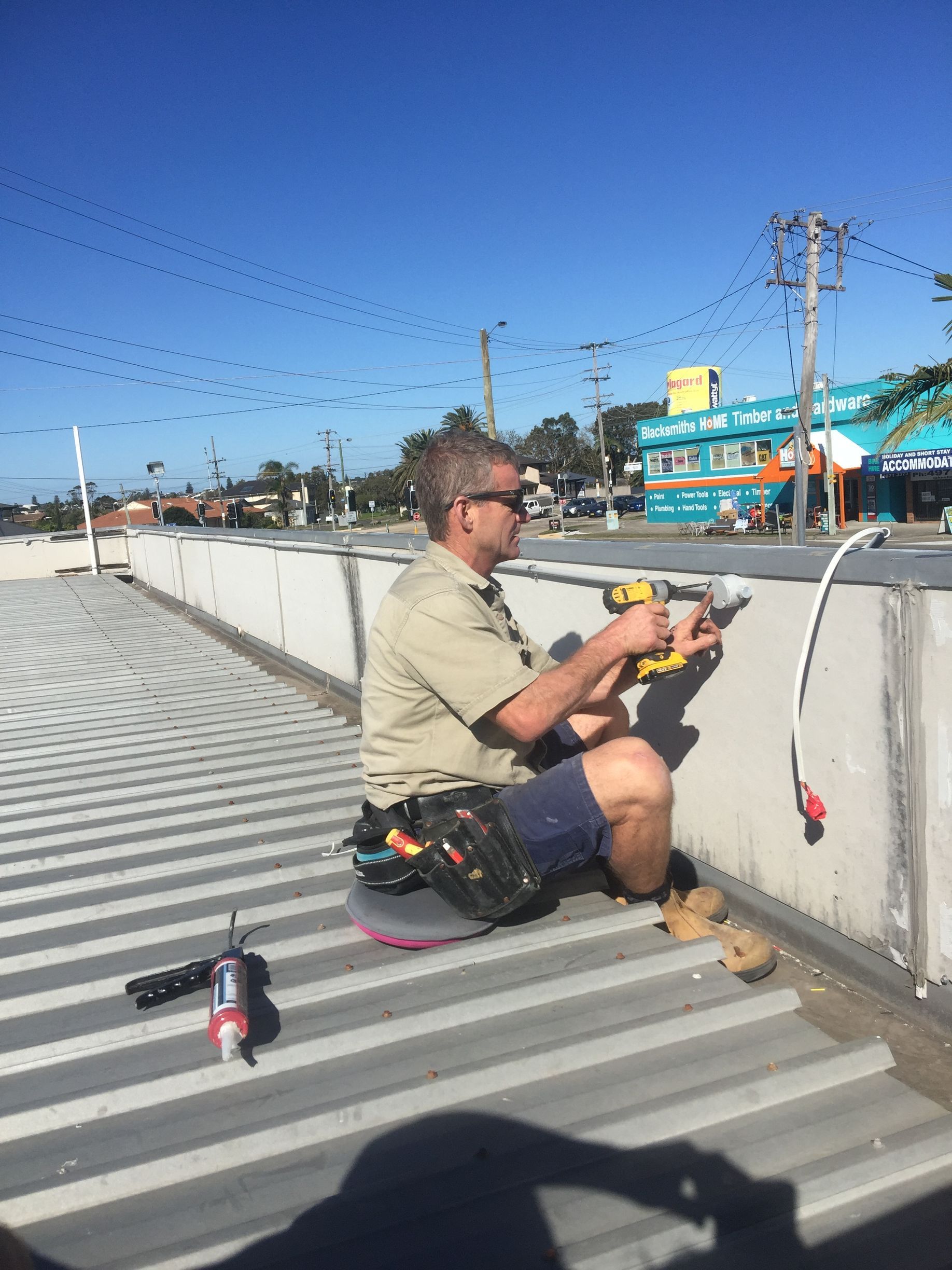 A person on a corrugated metal roof uses a power drill on a concrete wall — Channel Electrical In Swansea Heads, NSW