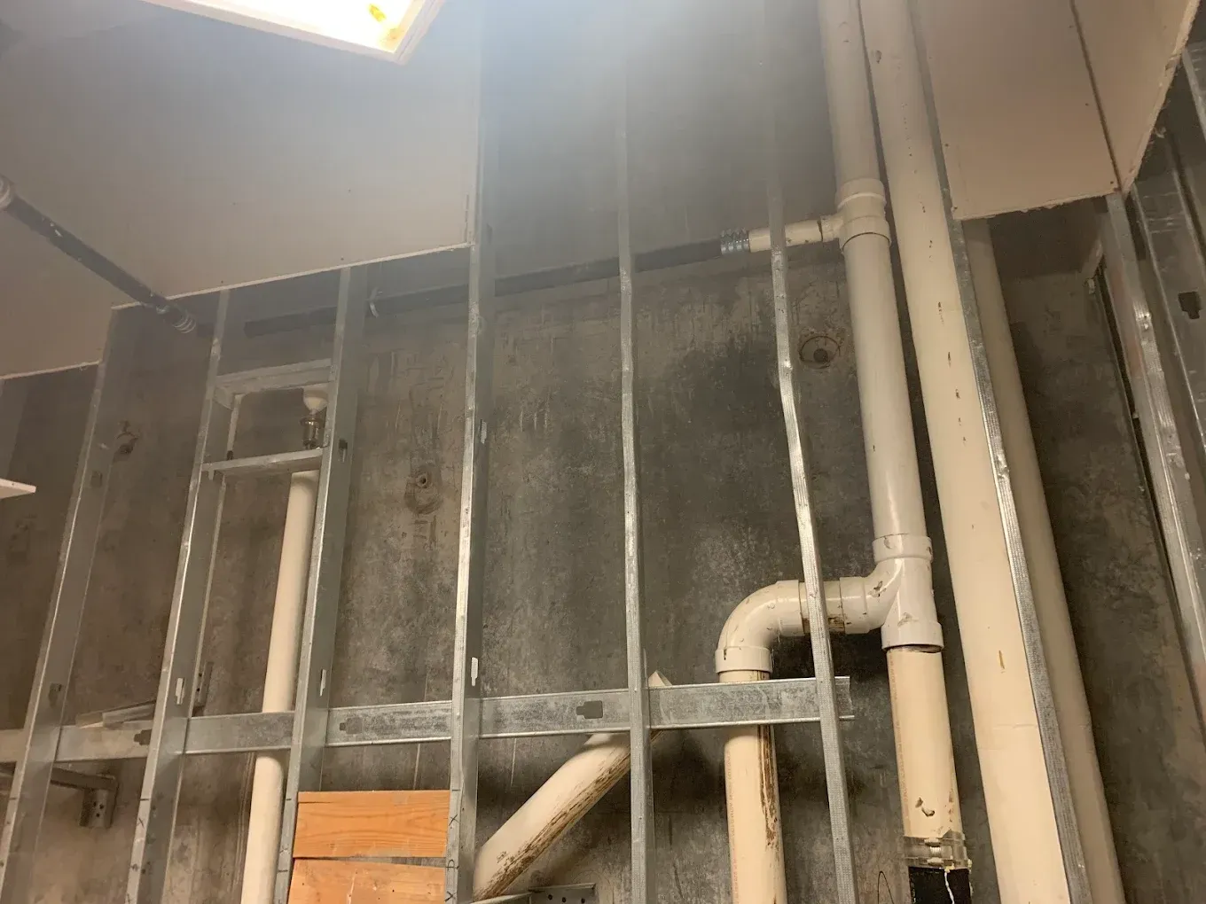 Interior view of metal studs and plumbing pipes against a dark wall. Construction or renovation setting.