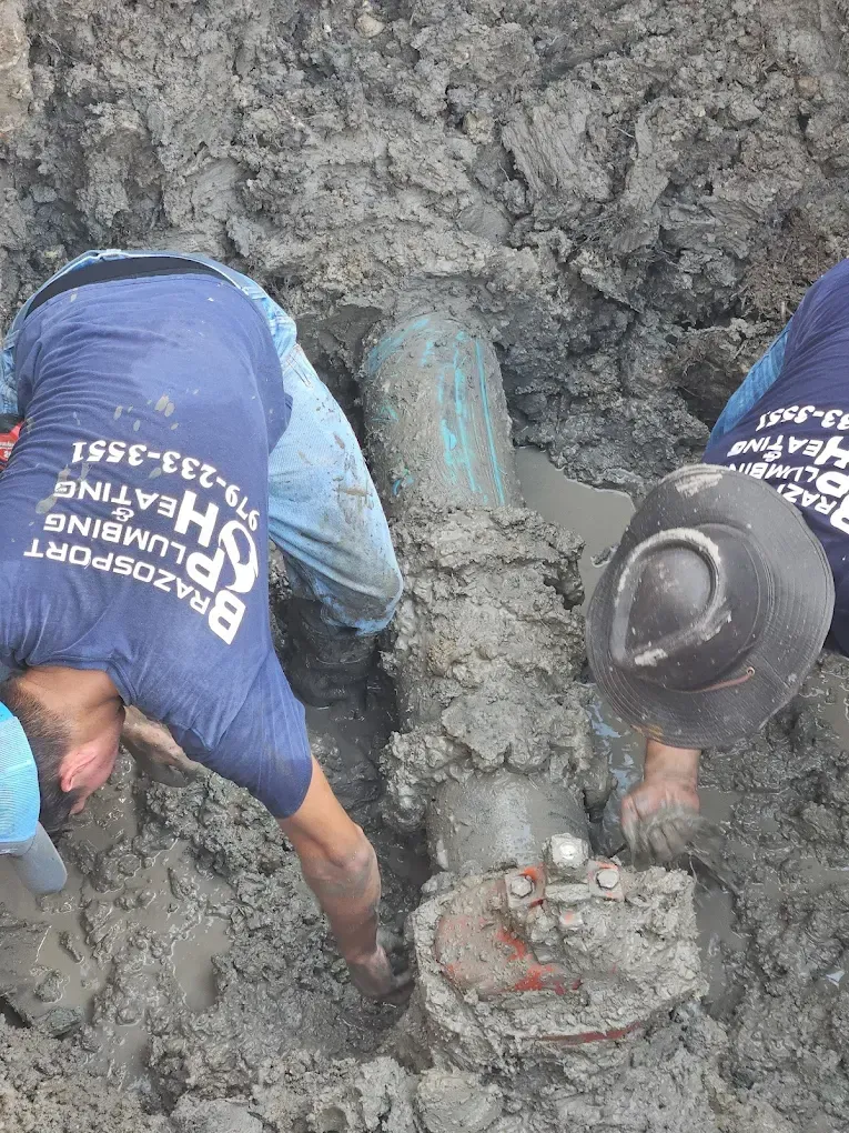 Two plumbers working on buried pipe in muddy trench.