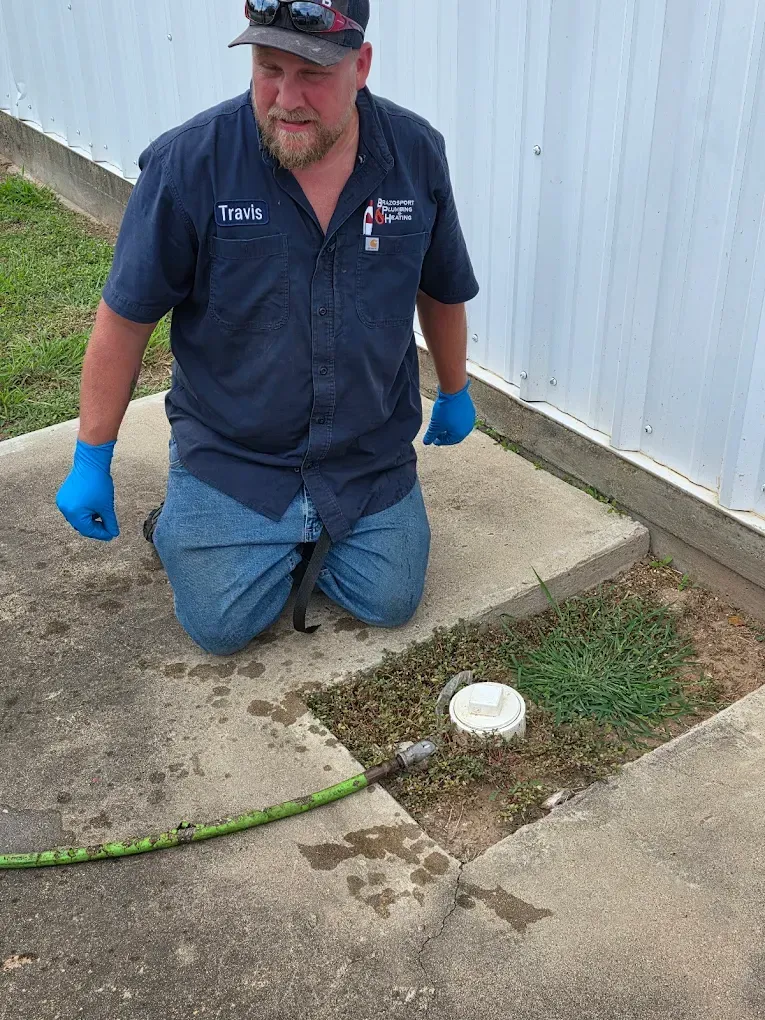 Man kneeling, cleaning a drain outside. He wears blue gloves and a work shirt. Concrete ground and white wall.