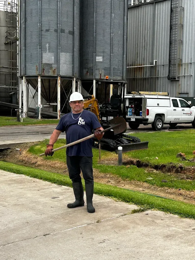 Man in hard hat and work clothes holding shovel near silos and truck.