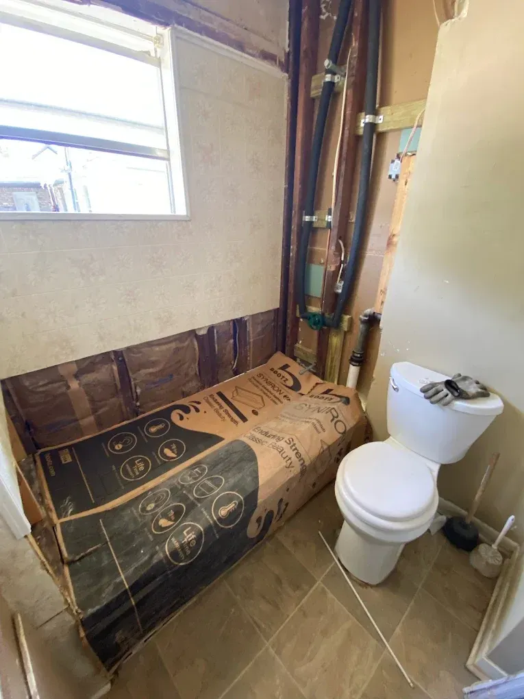 Bathroom with exposed wall, insulation covered, toilet, pipes. Light brown tile floor, white toilet.