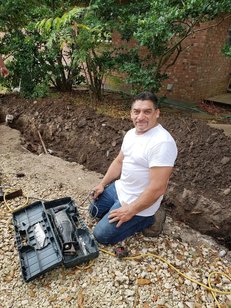 Man kneeling next to open tool case, trench in background. White shirt, jeans, outdoor setting.