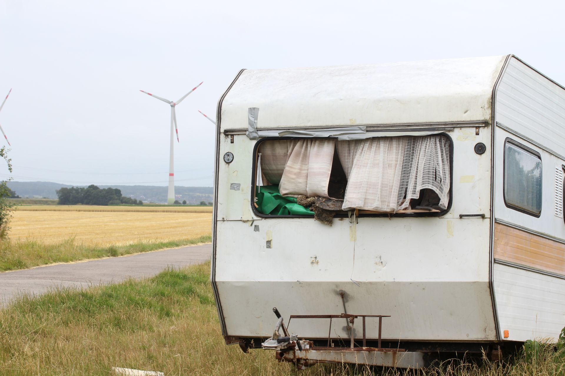 Old, weathered camper trailer in a field with a wheat field and windmills in the background.