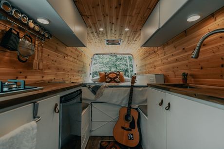 Cozy camper van interior with wood paneling, bed, guitar, and kitchen area.