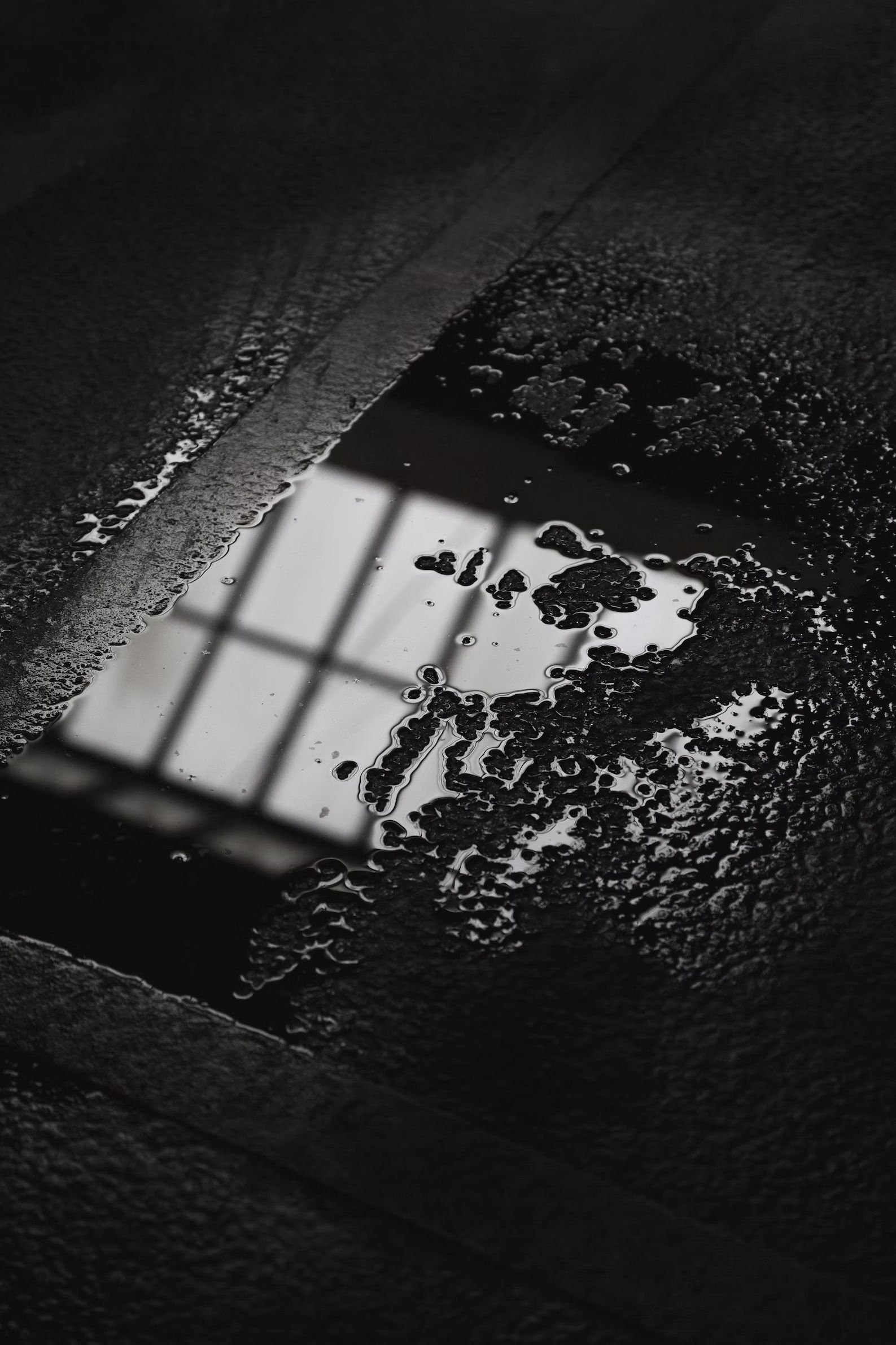 Puddle on dark pavement reflects a window with a grid pattern, in grayscale.