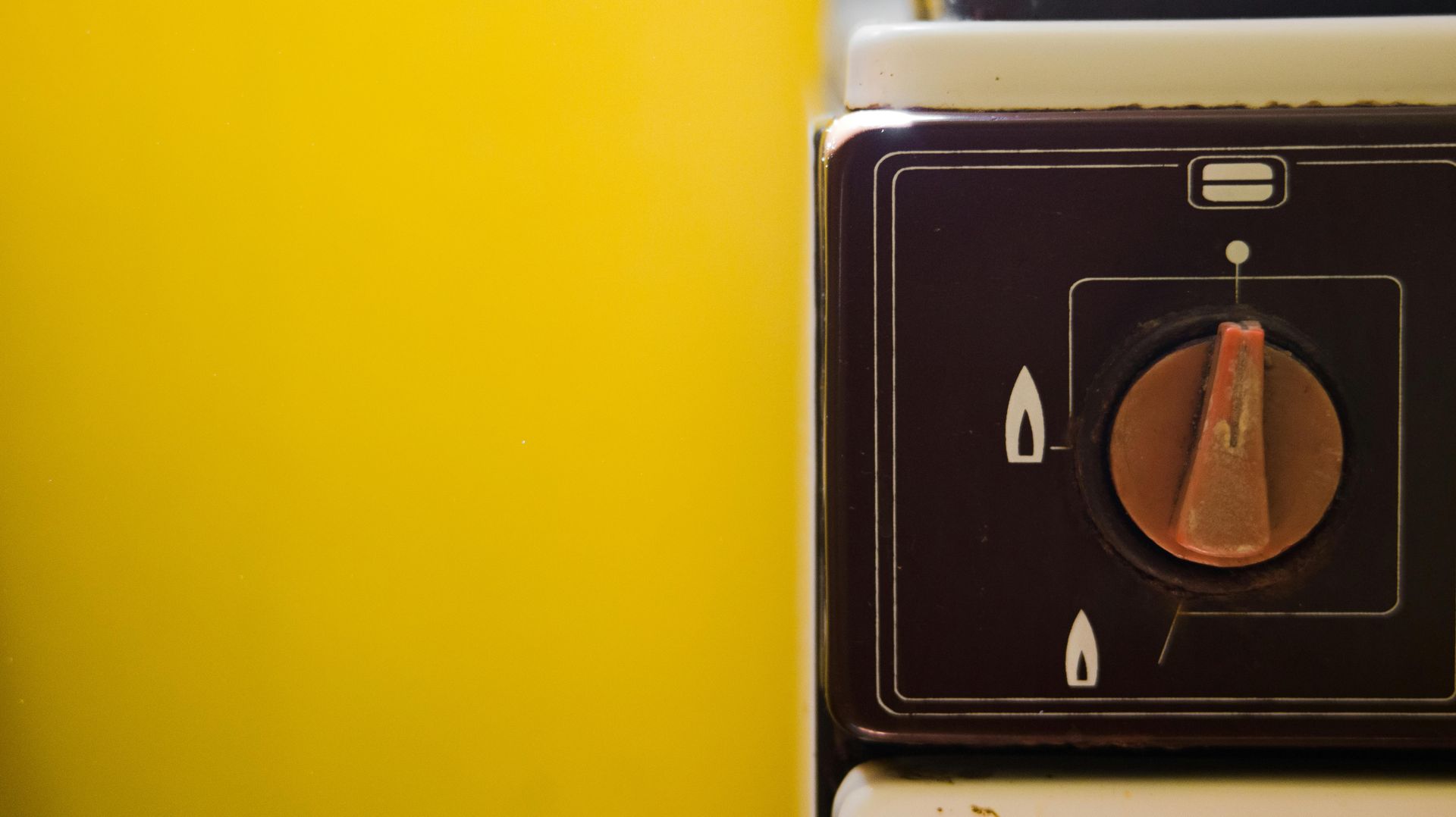 Brown oven control knob on a dark panel with flame icons, against a yellow wall.