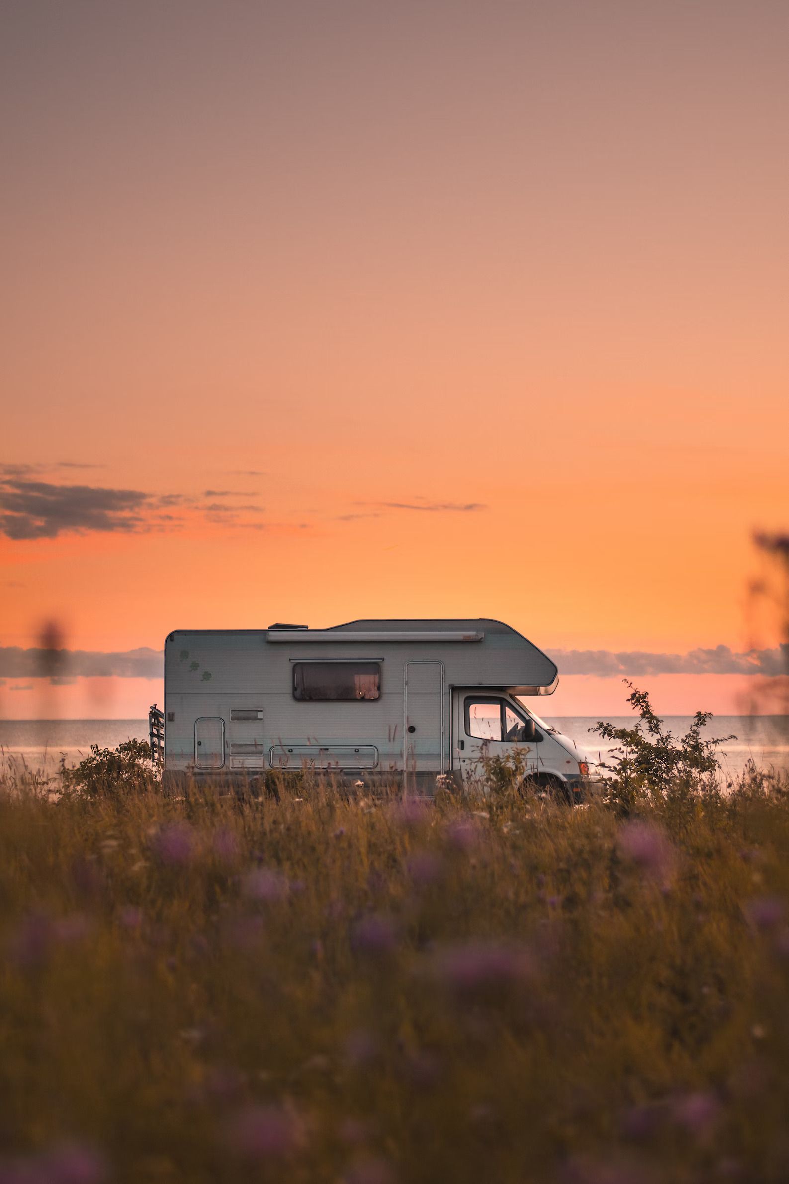 RV parked on a grassy shore at sunset, with orange and pink sky.
