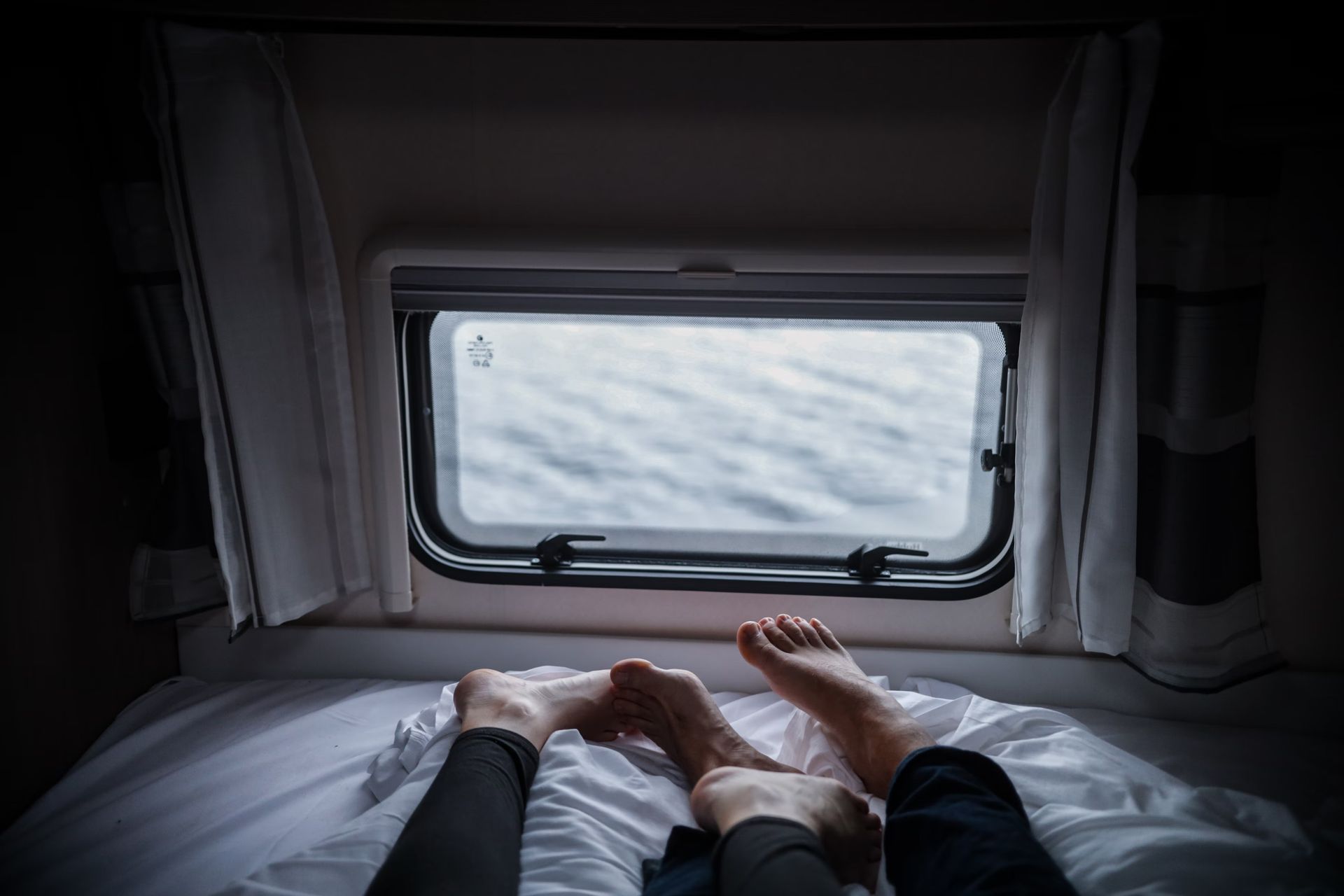 Two pairs of feet on a bed in front of a window with a view of water.