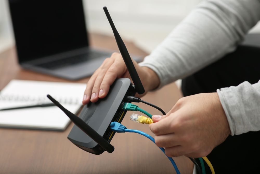 Person plugging colored cables into a black wireless router with antennas on a desk