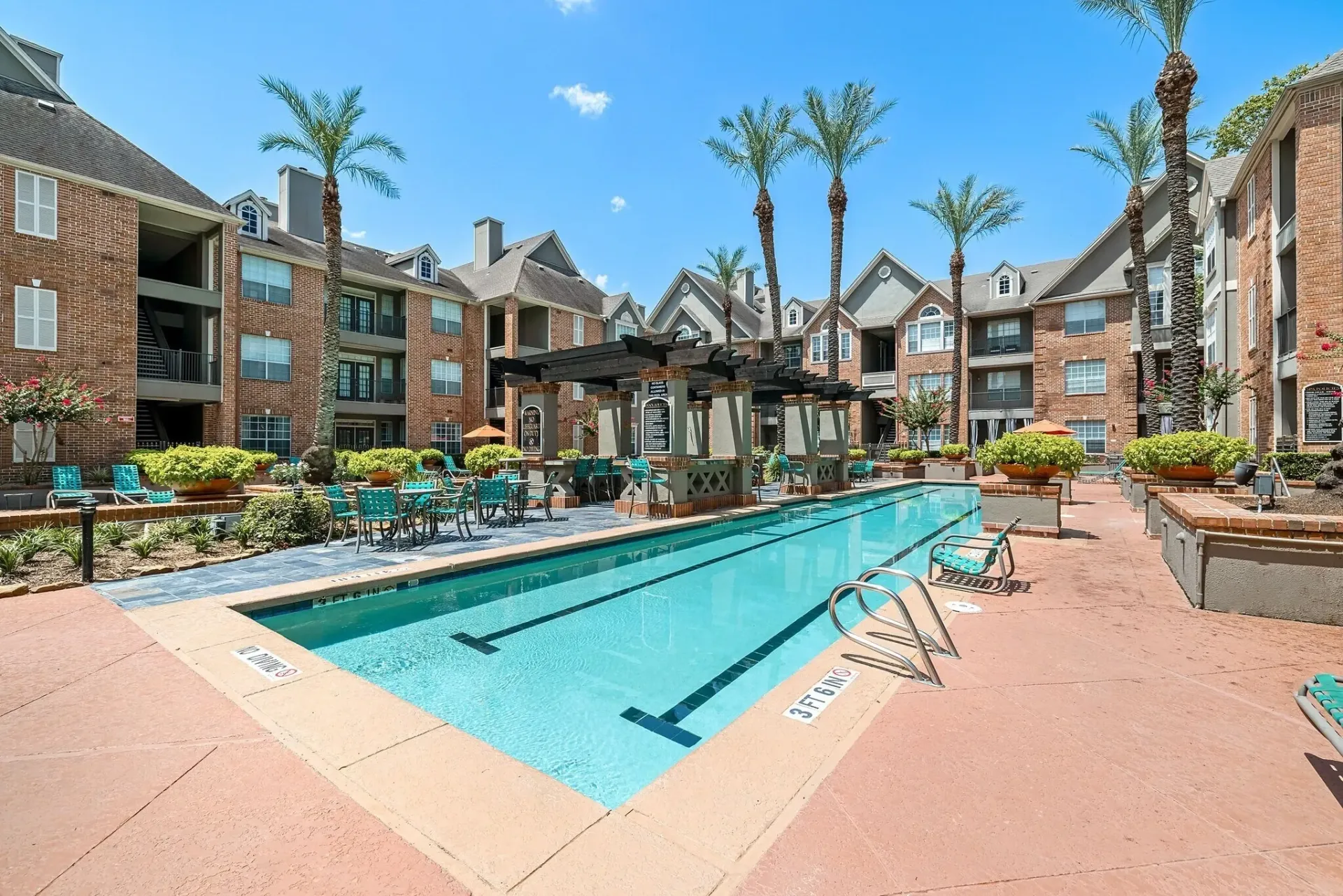 Resort-style swimming pool with lounge chairs and tables in a courtyard setting with apartment buildings in the background.