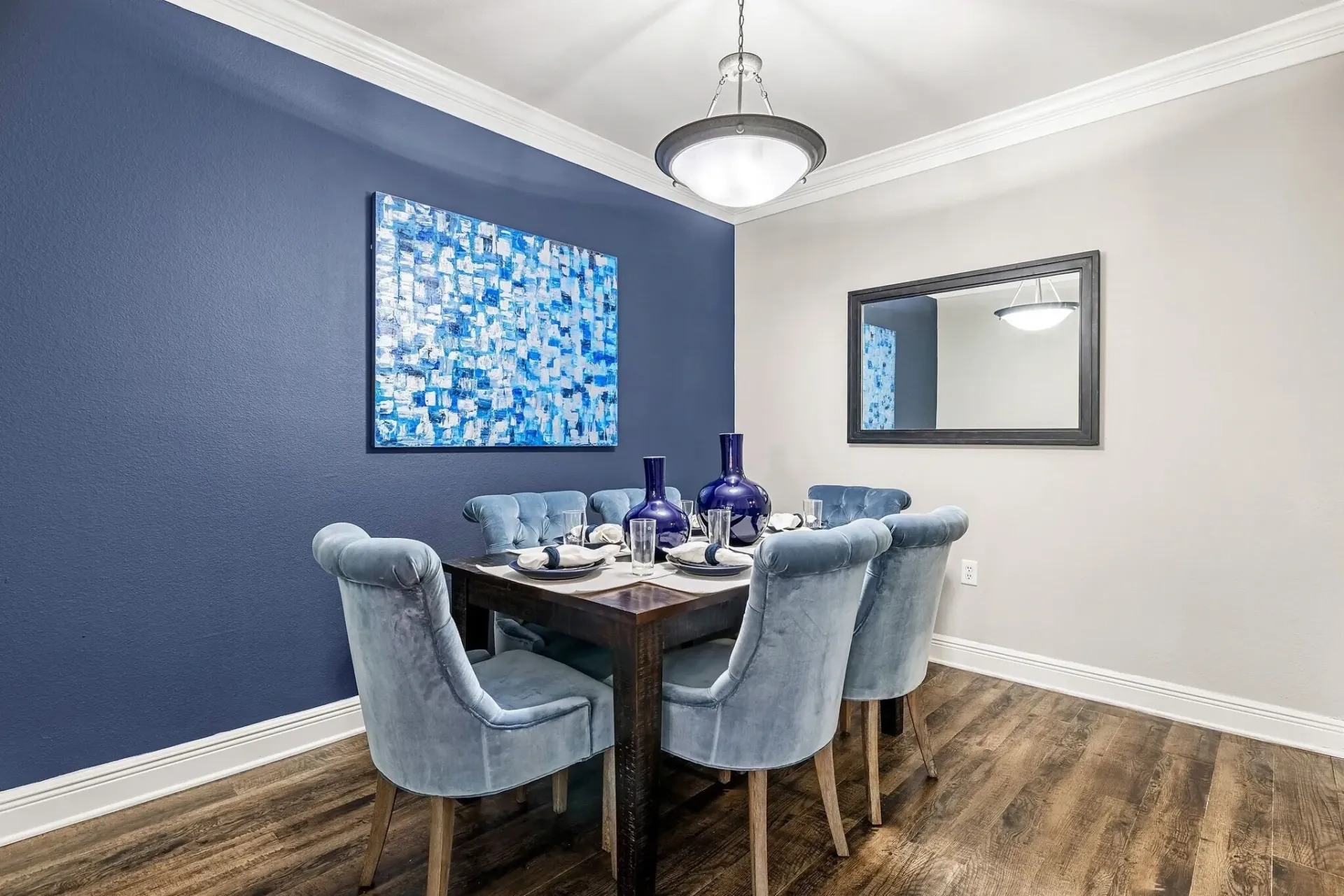 Dining room with a wooden table and blue velvet chairs, abstract art on wall.