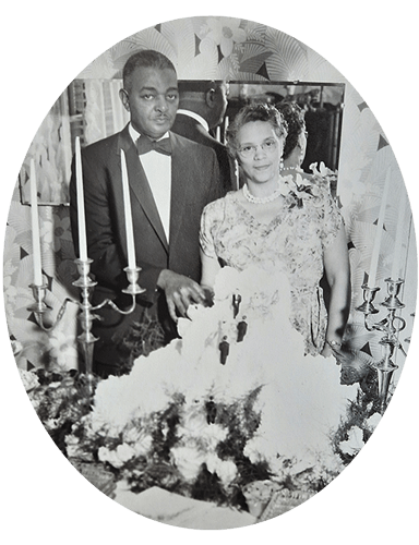 A formally dressed couple stands by their wedding cake. The man wears a tuxedo, the woman a floral dress.