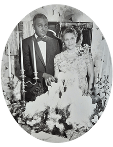 A formally dressed couple stands by their wedding cake. The man wears a tuxedo, the woman a floral dress.