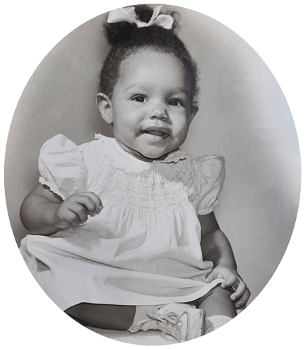 Young child in a white dress, sitting and smiling, with a bow in their hair, in a studio setting.